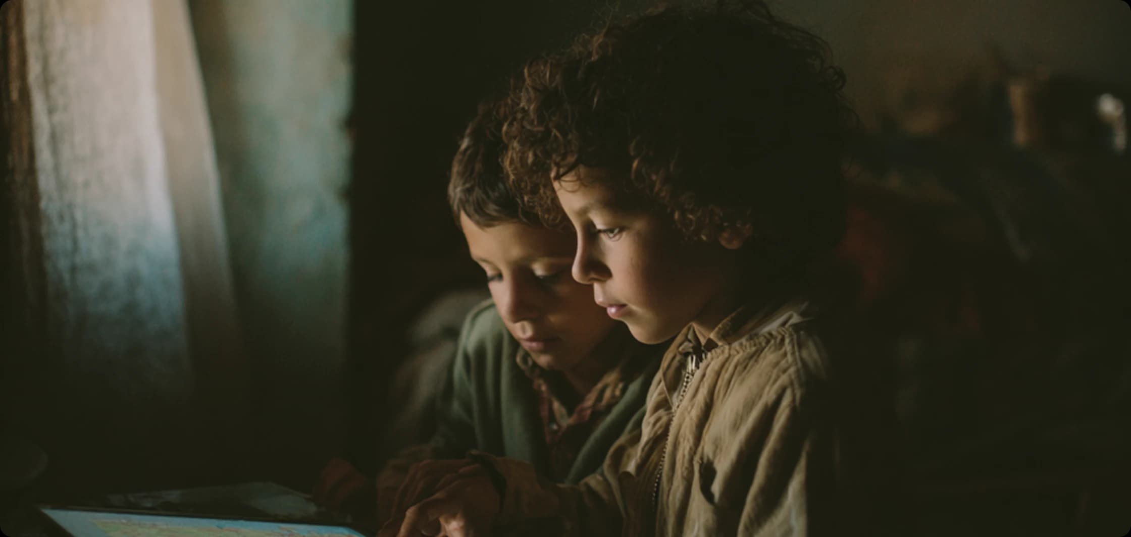Two young children in a dimly lit room, their faces illuminated by the cool blue light of a tablet screen they are sharing.