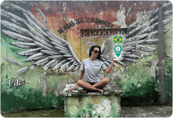 A woman in a VayaPin t-shirt sits cross-legged in front of a colorful mural of angel wings on a weathered wall in Brazil